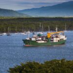 A cargo ship steaming down the Trinity Inlet shipping channel at Cairns past moored yachts, with green rainforest hills behind