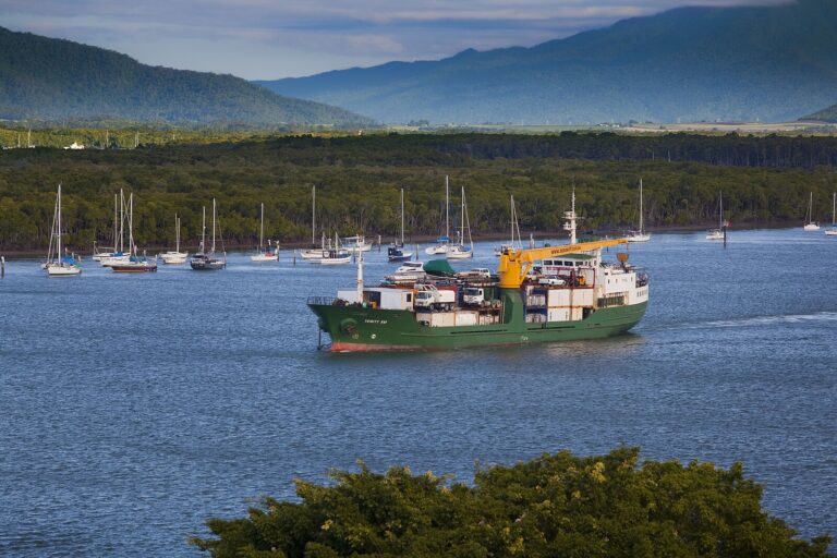 A cargo ship steaming down the Trinity Inlet shipping channel at Cairns past moored yachts, with green rainforest hills behind