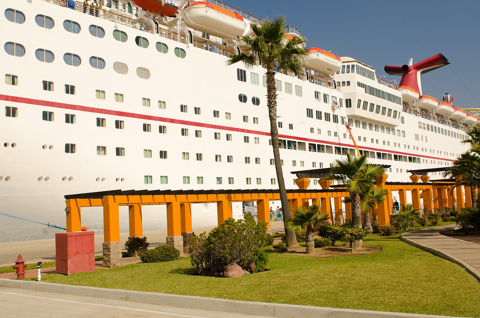 Carnival cruise ship docked beside an orange pergola, palm trees and green lawn under a blue sky