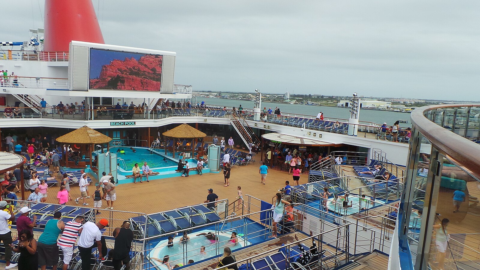 Crowded lido deck and Beach Pool on Carnival Sunshine with a big outdoor screen and blue loungers