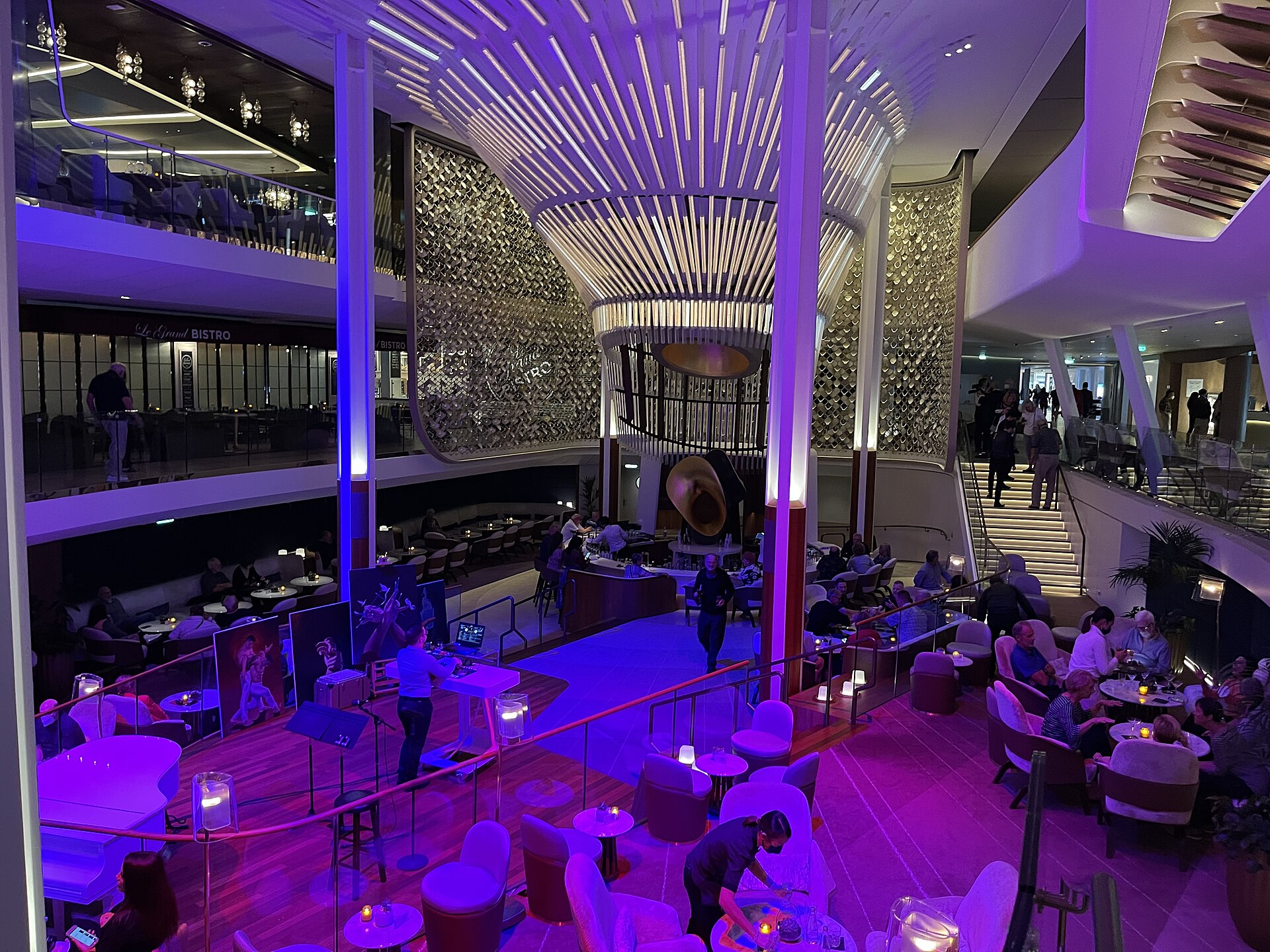 The Grand Plaza atrium on Celebrity Apex lit in purple and blue with a glowing chandelier