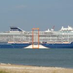 Celebrity Beyond cruise ship with a navy blue hull and orange Magic Carpet platform, seen from a sandy beach