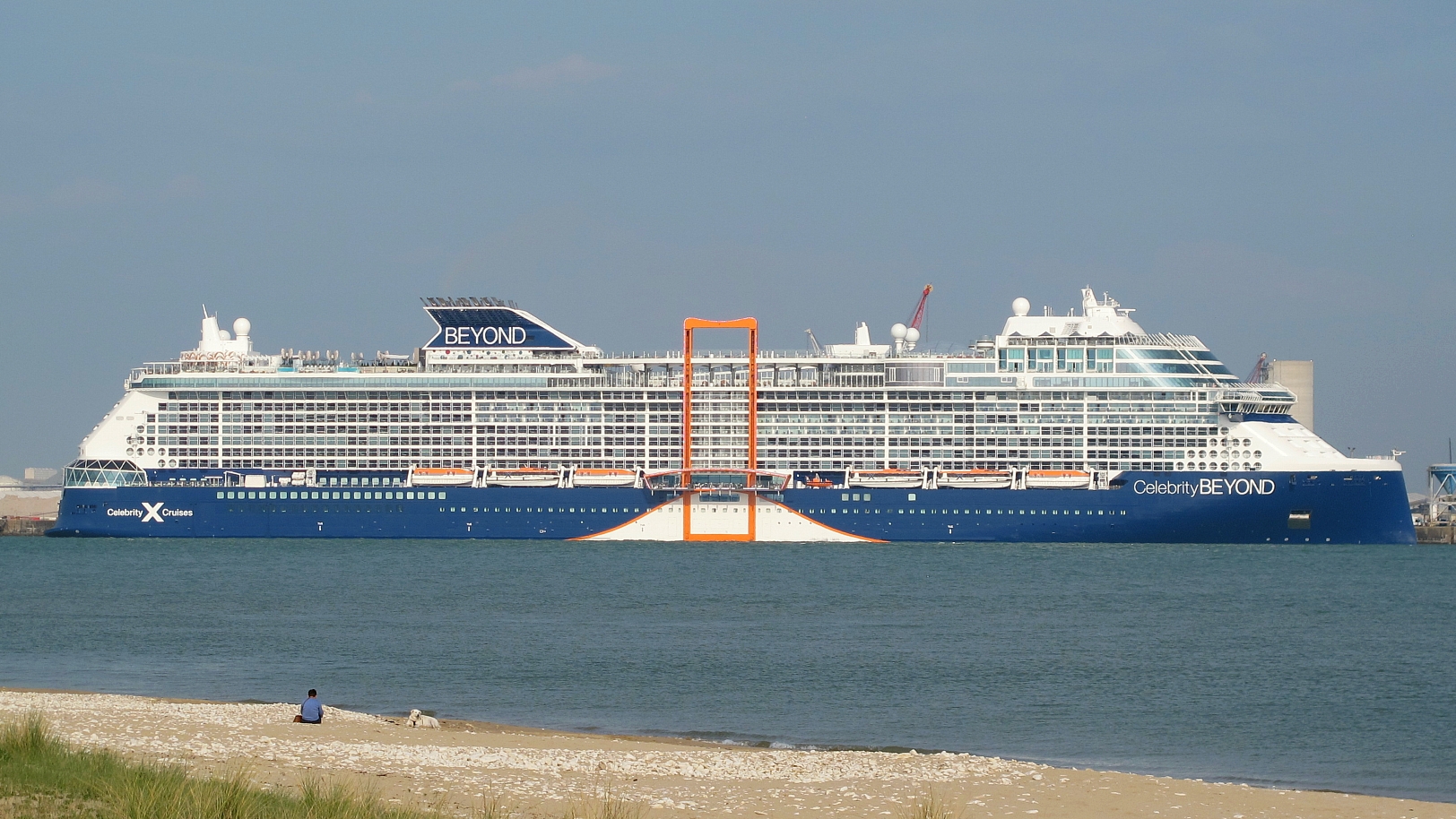 Celebrity Beyond cruise ship with a navy blue hull and orange Magic Carpet platform, seen from a sandy beach