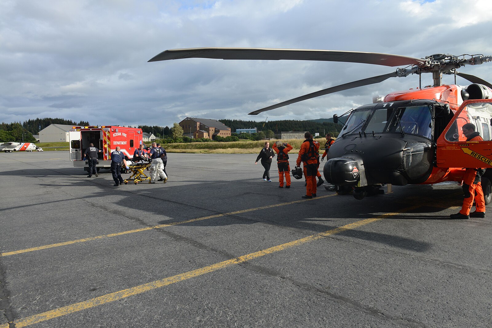An orange Coast Guard helicopter on the tarmac at Kodiak, Alaska as crews wheel a stretcher to a waiting red fire department ambulance