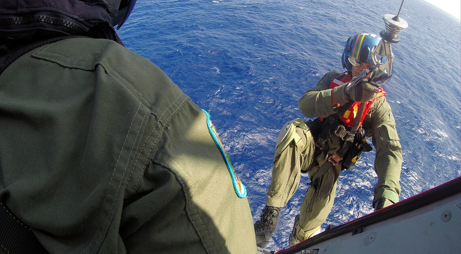 A US Coast Guard rescue swimmer is lowered on a hoist cable from a helicopter towards a cruise ship over deep blue ocean