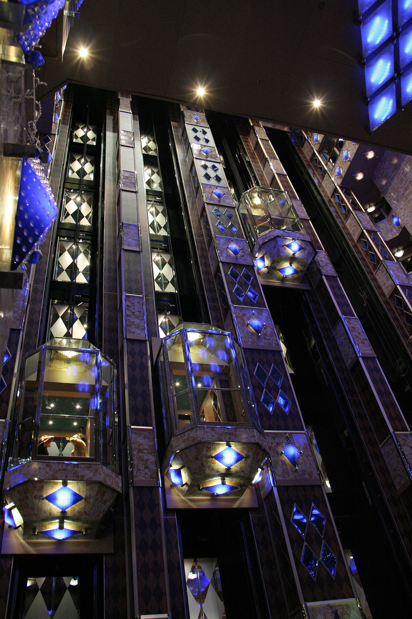 Brightly lit blue and gold glass elevators rising through the atrium of a Costa cruise ship
