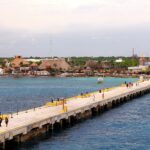 Cruise passengers walking along the long pier at Costa Maya, Mexico, over turquoise water toward the palm-thatched port village