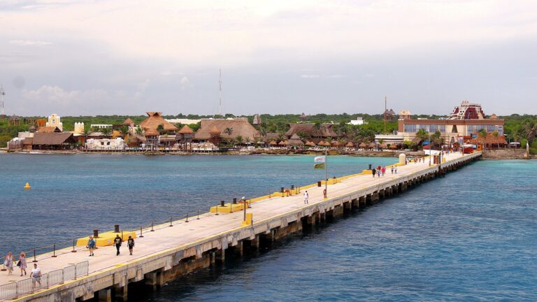 Cruise passengers walking along the long pier at Costa Maya, Mexico, over turquoise water toward the palm-thatched port village
