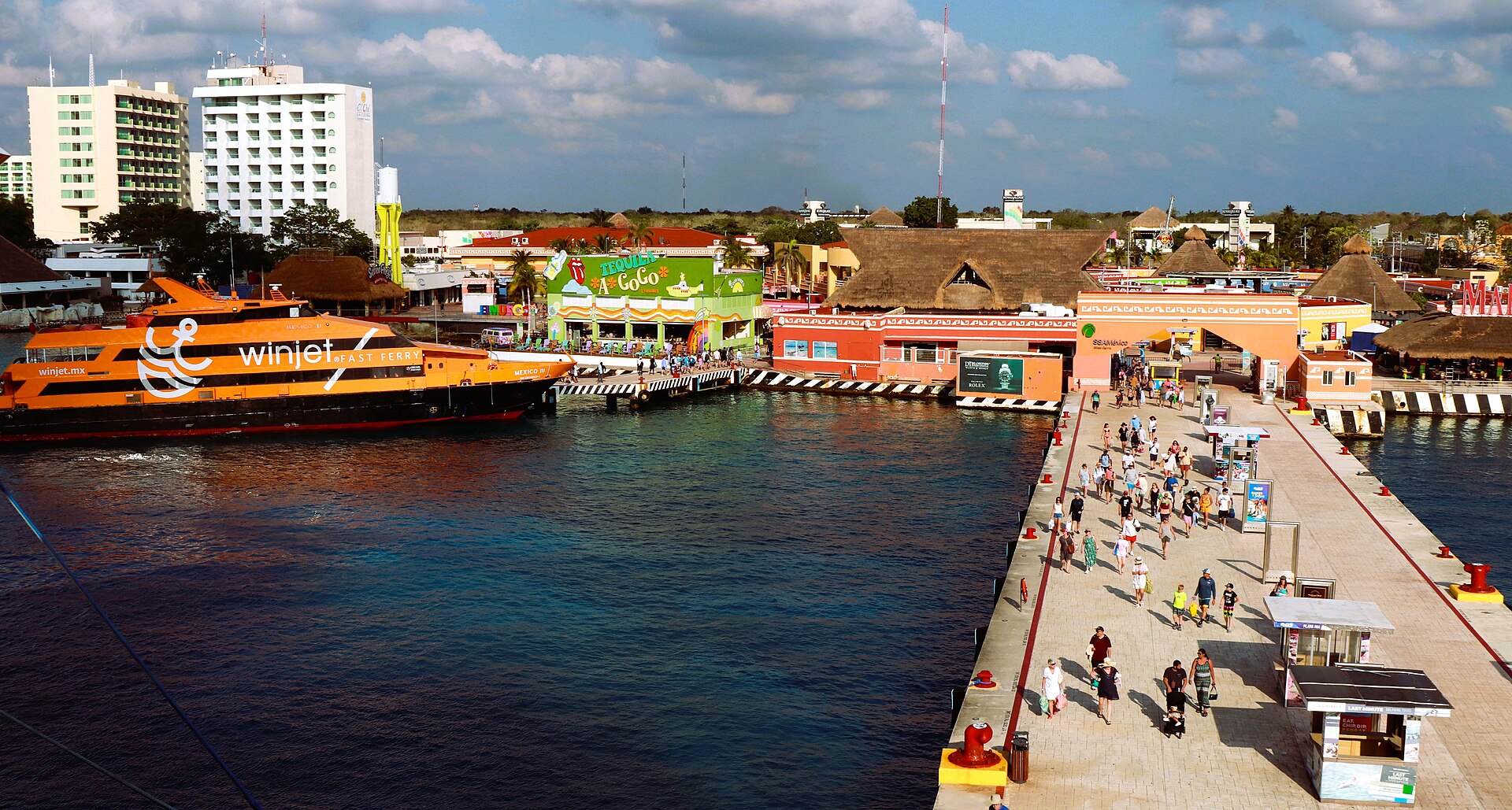 Cruise passengers walking along a pier toward colourful shops and restaurants on the Cozumel waterfront, beside an orange ferry