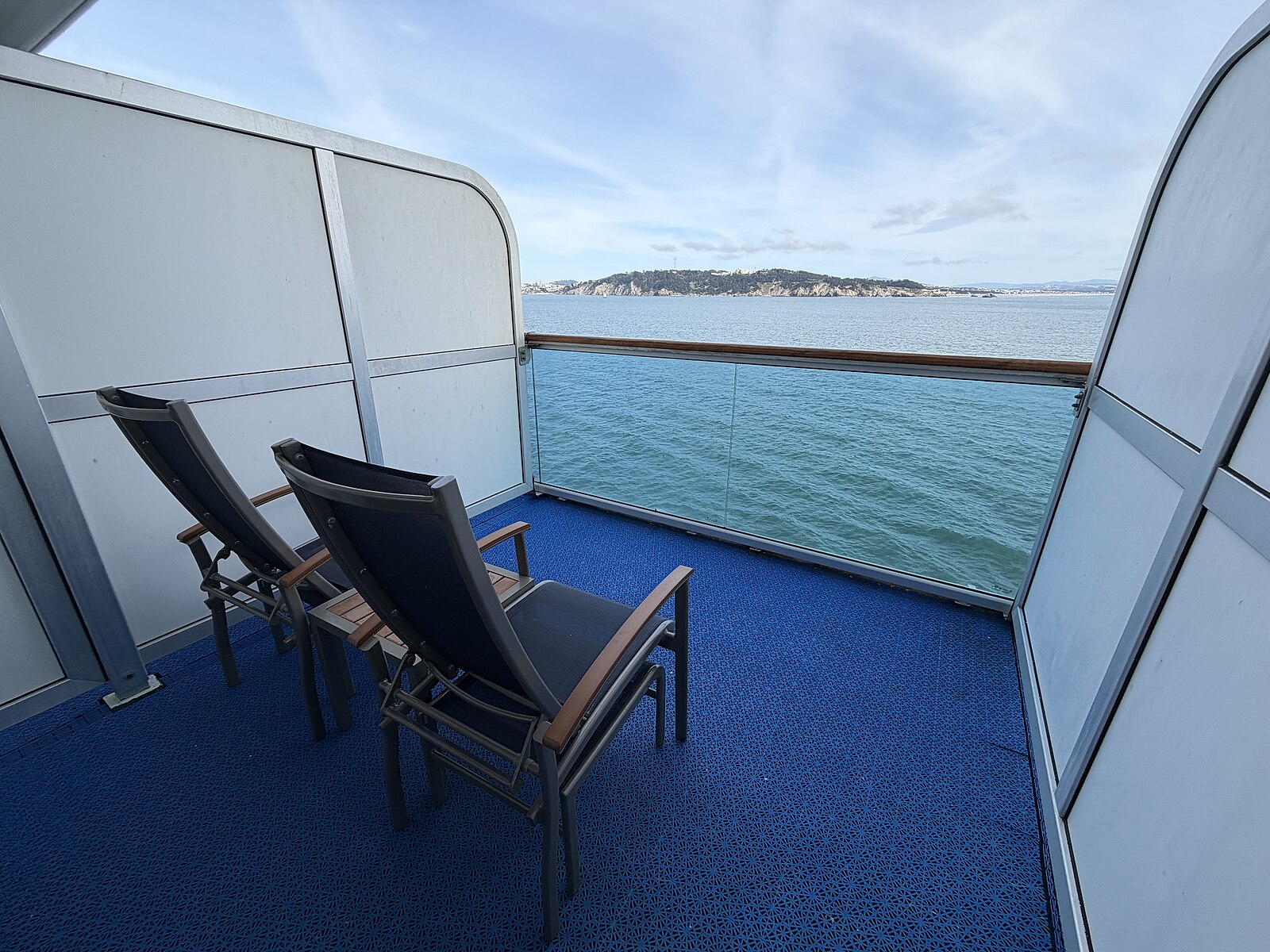 Cruise ship cabin balcony with blue carpet, two loungers and a glass rail above a teal sea