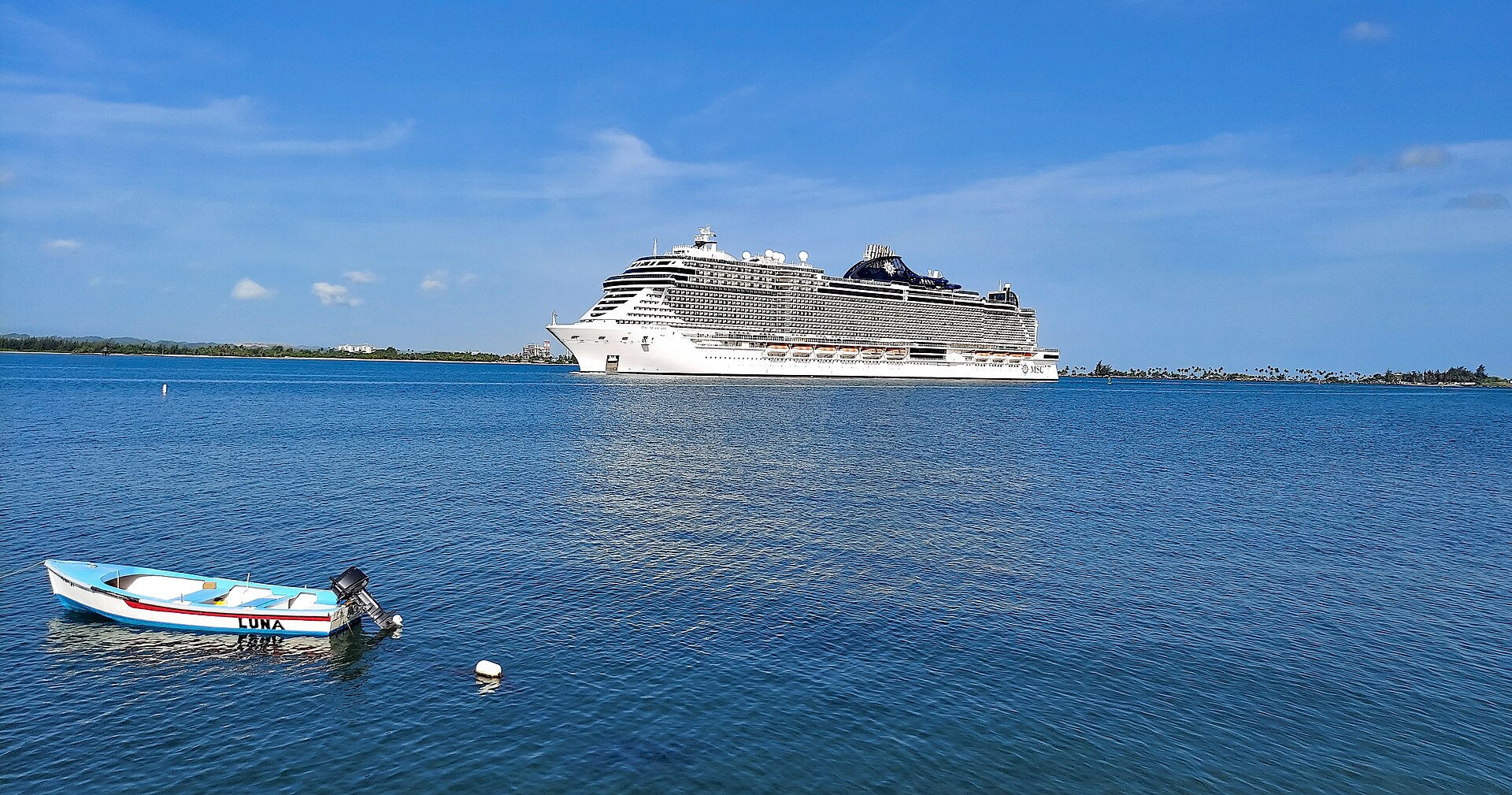 Large cruise ship entering San Juan Harbor, Puerto Rico, under a bright blue sky with a small blue fishing boat in the foreground