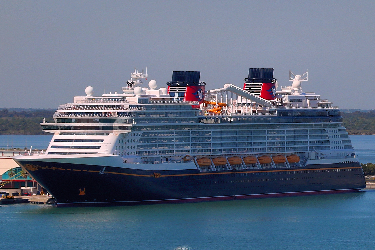 Disney Wish with its navy hull, red funnels and orange lifeboats docked at Port Canaveral on blue water