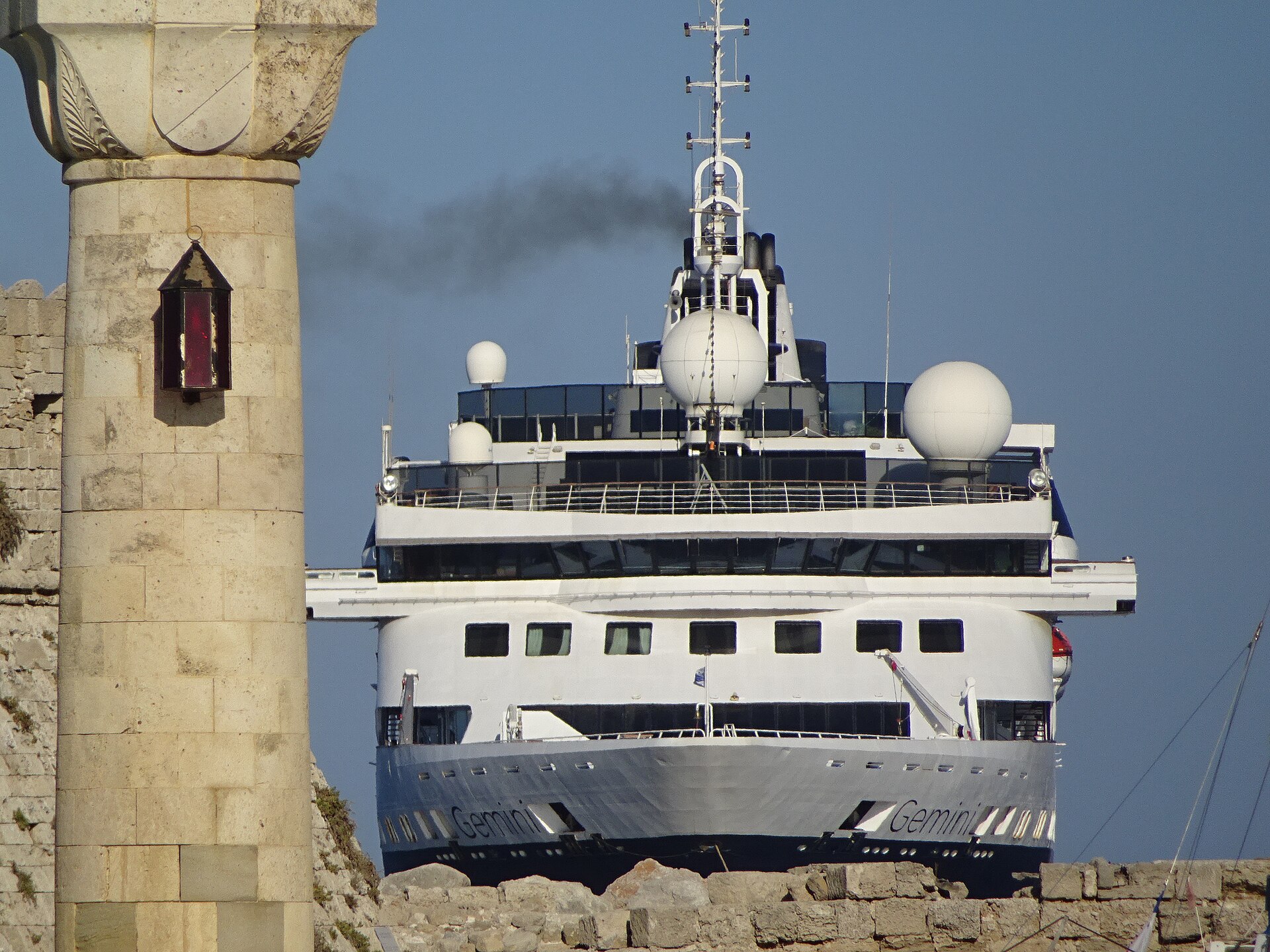 The bow of the cruise ship Gemini rising behind the stone Agios Nikolaos fortress in Rhodes under a blue sky