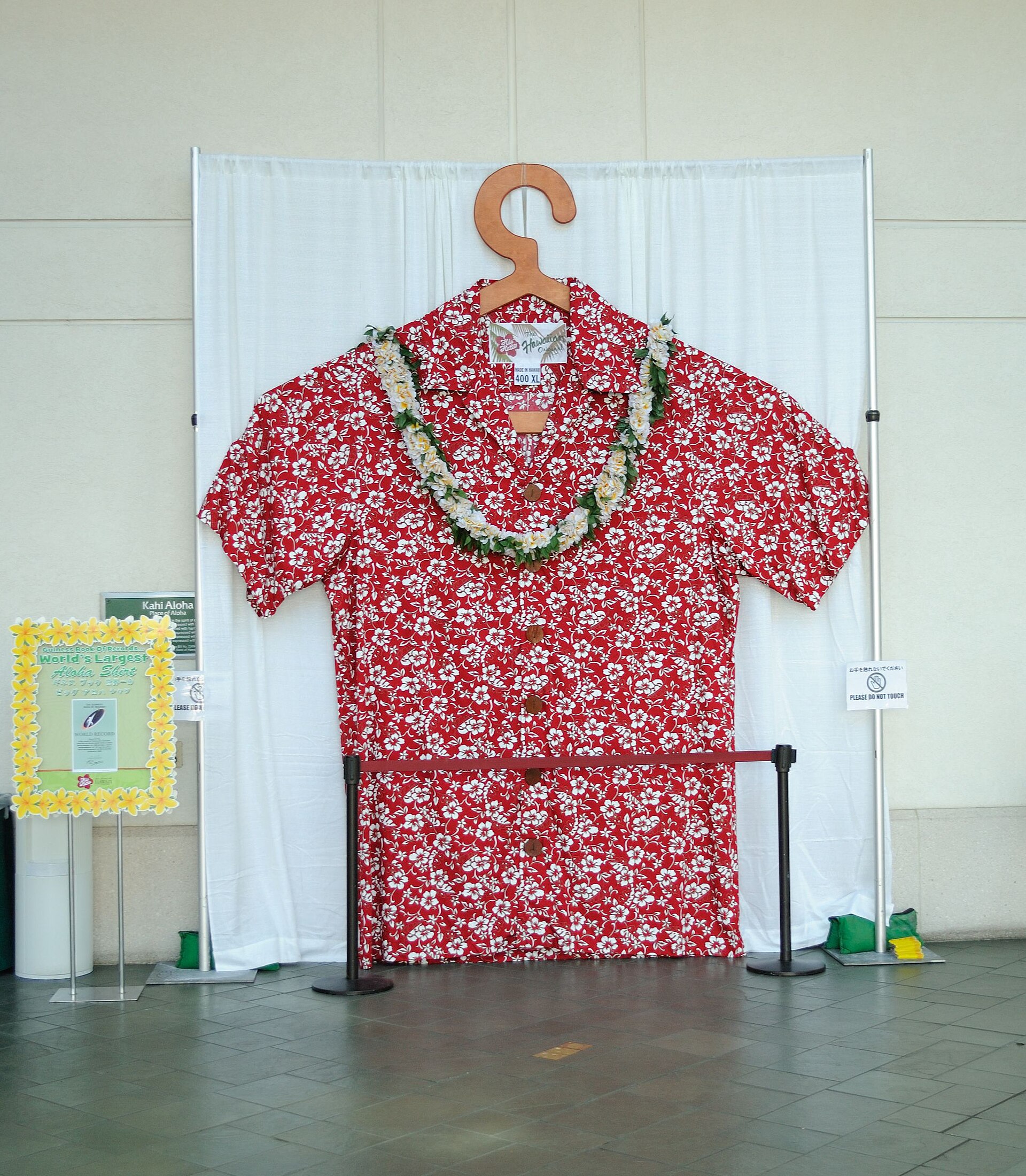 A giant red aloha shirt with a white hibiscus print and a flower lei, displayed on a huge wooden hanger in Honolulu