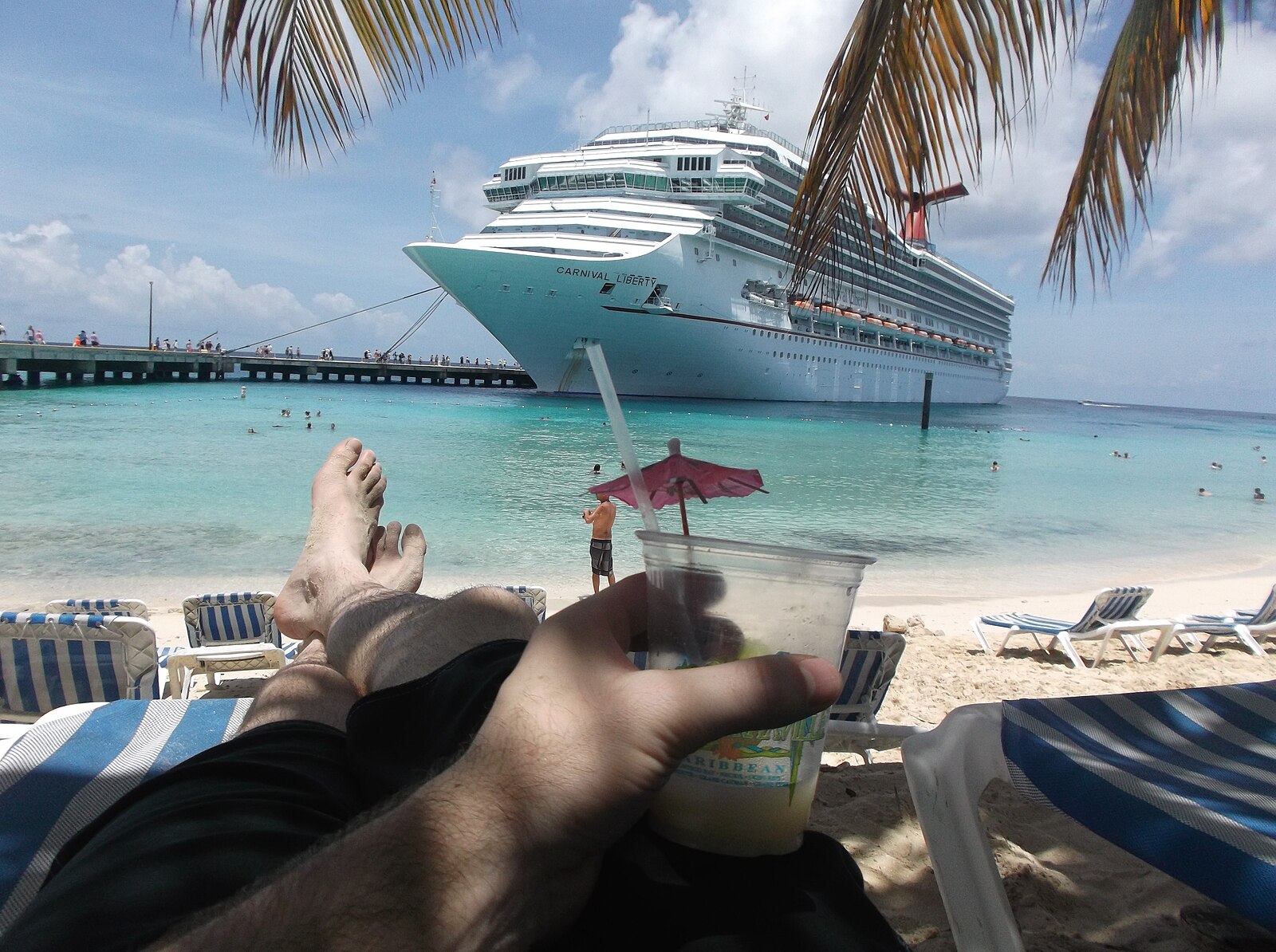 View from a beach lounger at Grand Turk Cruise Center with a cocktail, palm fronds and a Carnival cruise ship at the pier