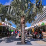Brightly painted orange, green and pink vendor stalls under palm trees beside a docked cruise ship at the Nassau Cruise Port