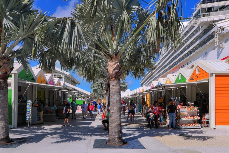 Brightly painted orange, green and pink vendor stalls under palm trees beside a docked cruise ship at the Nassau Cruise Port