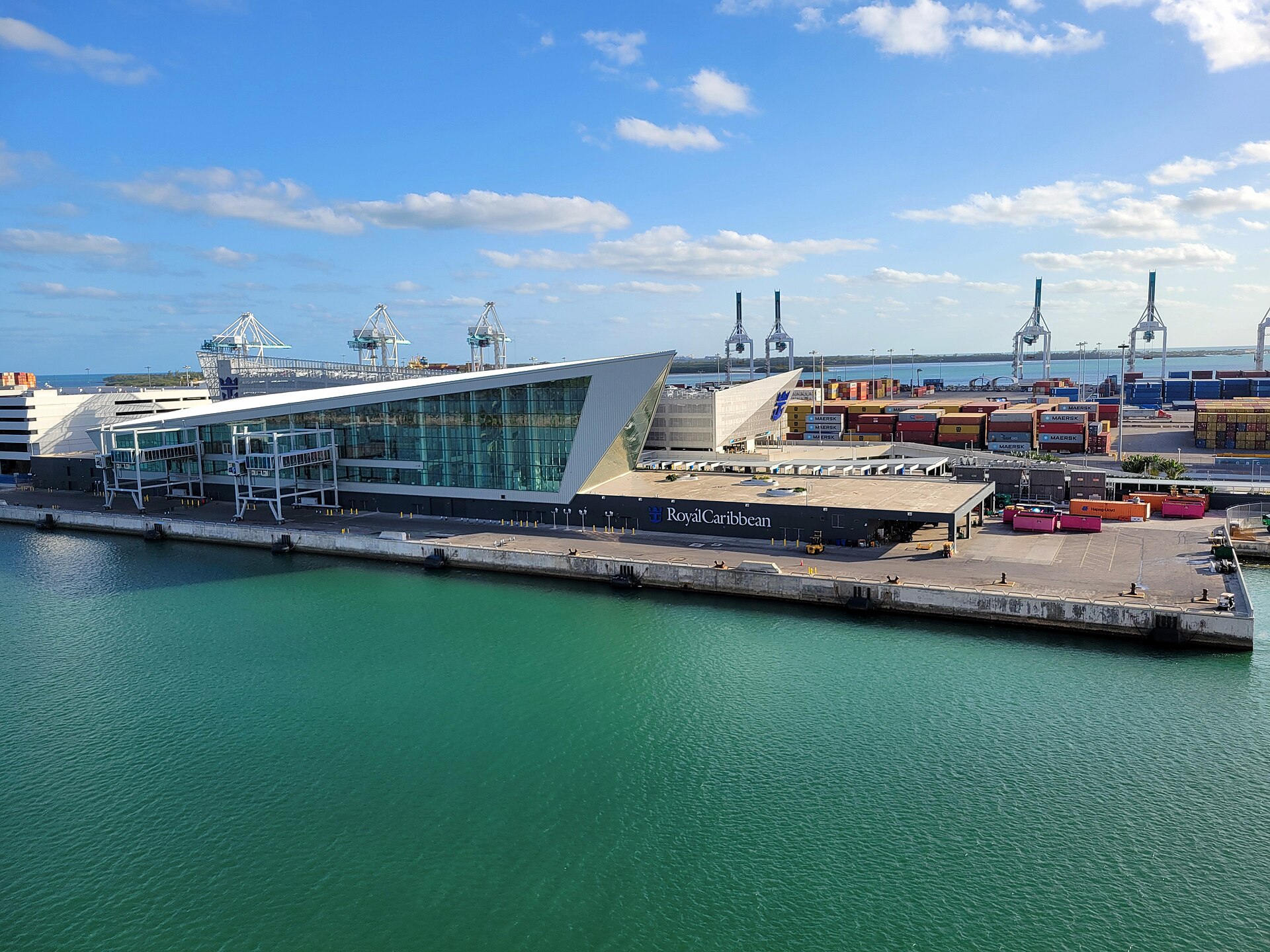 Royal Caribbean's glass-fronted Terminal A at PortMiami beside green-blue water under a blue sky with white clouds