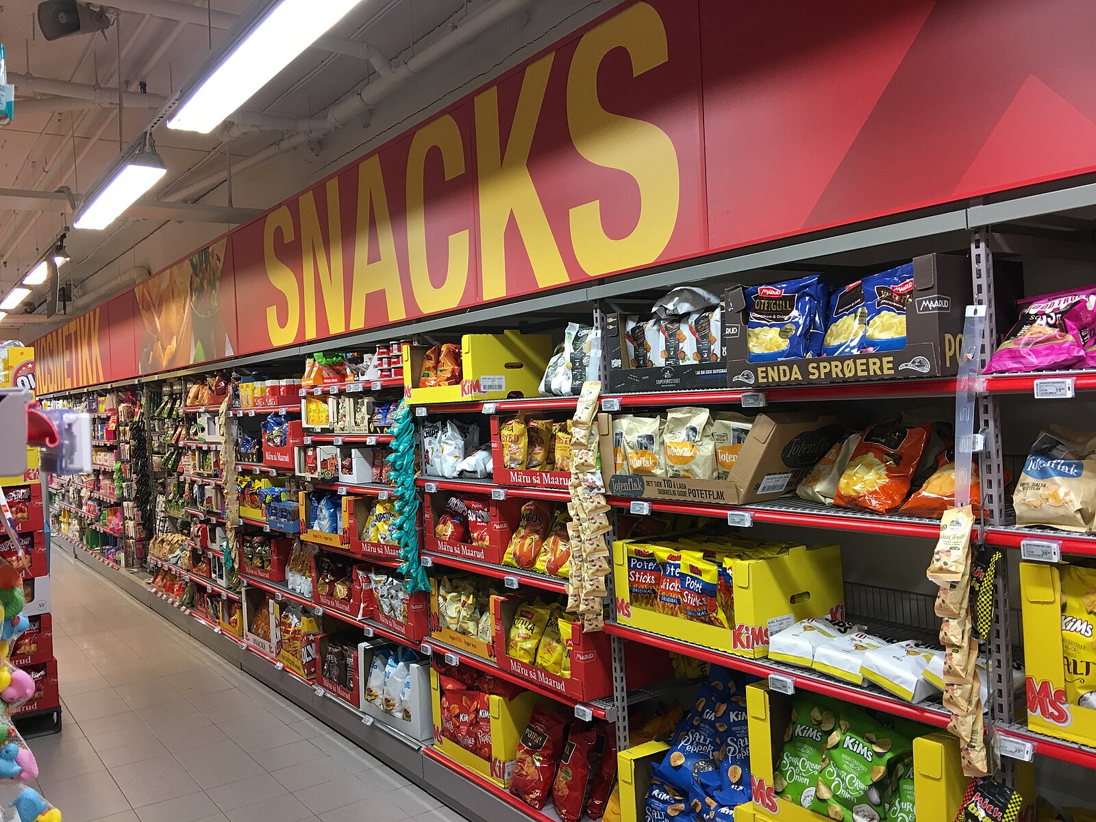 Supermarket snack aisle stacked with colorful factory-sealed bags of chips under a big red SNACKS sign