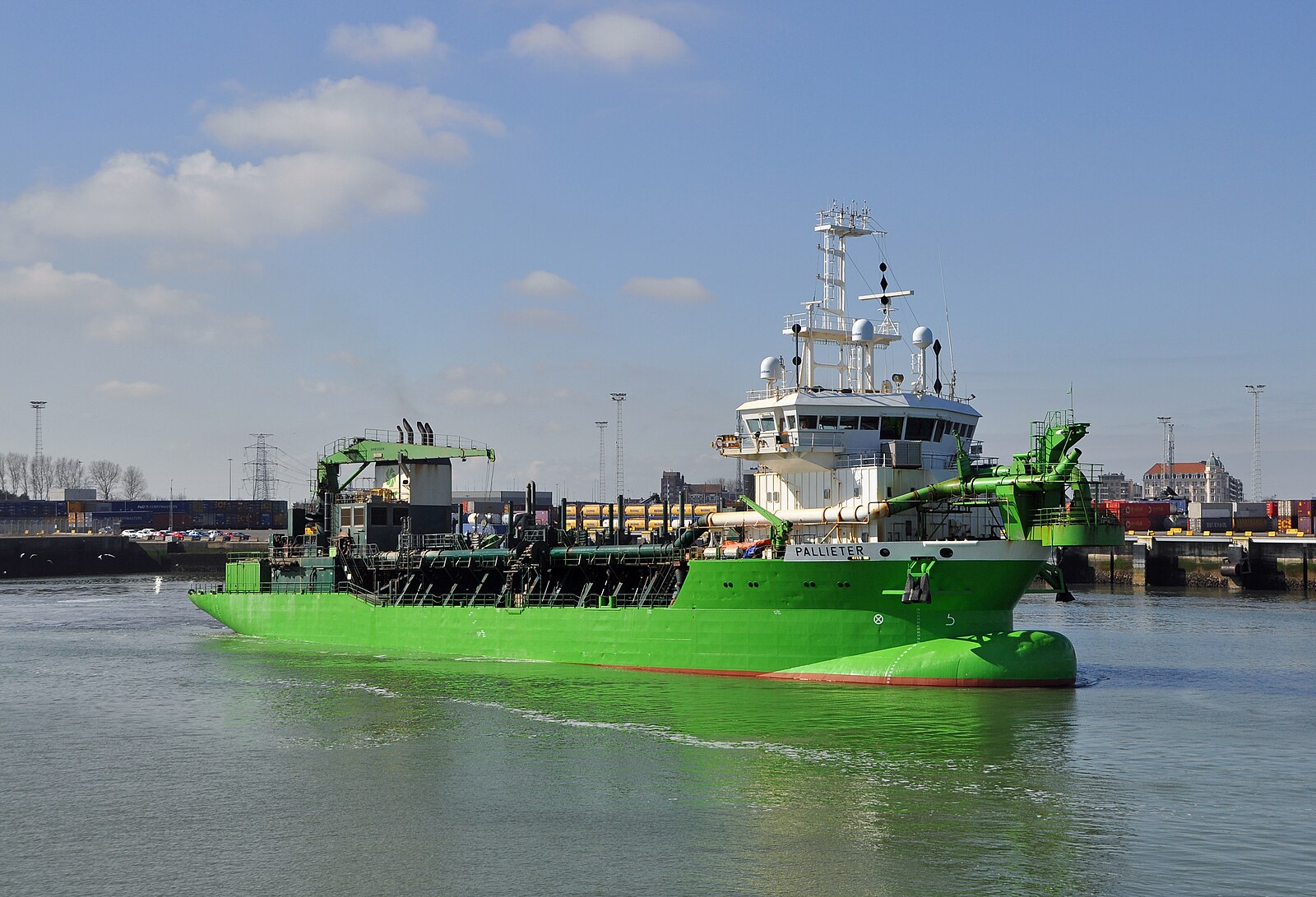 A bright green trailing suction hopper dredger under way in a harbour under a blue sky