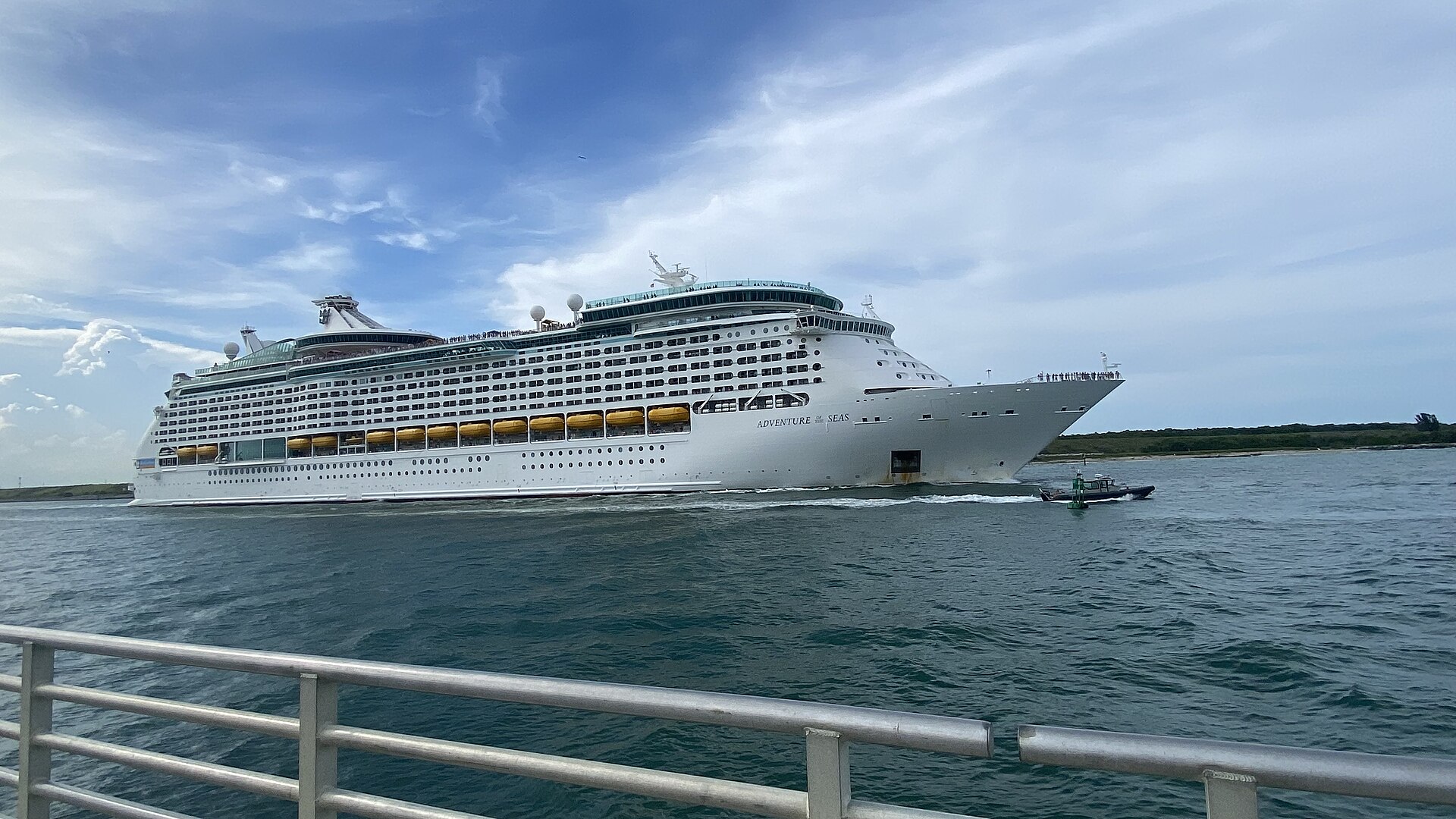 Royal Caribbean's Adventure of the Seas sailing out of Port Canaveral past a waterfront railing, with a small boat alongside
