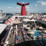 Sunbathers on loungers around the blue Lido Deck pool of Carnival Fantasy under the red whale-tail funnel, with Nassau and Carnival Pride behind