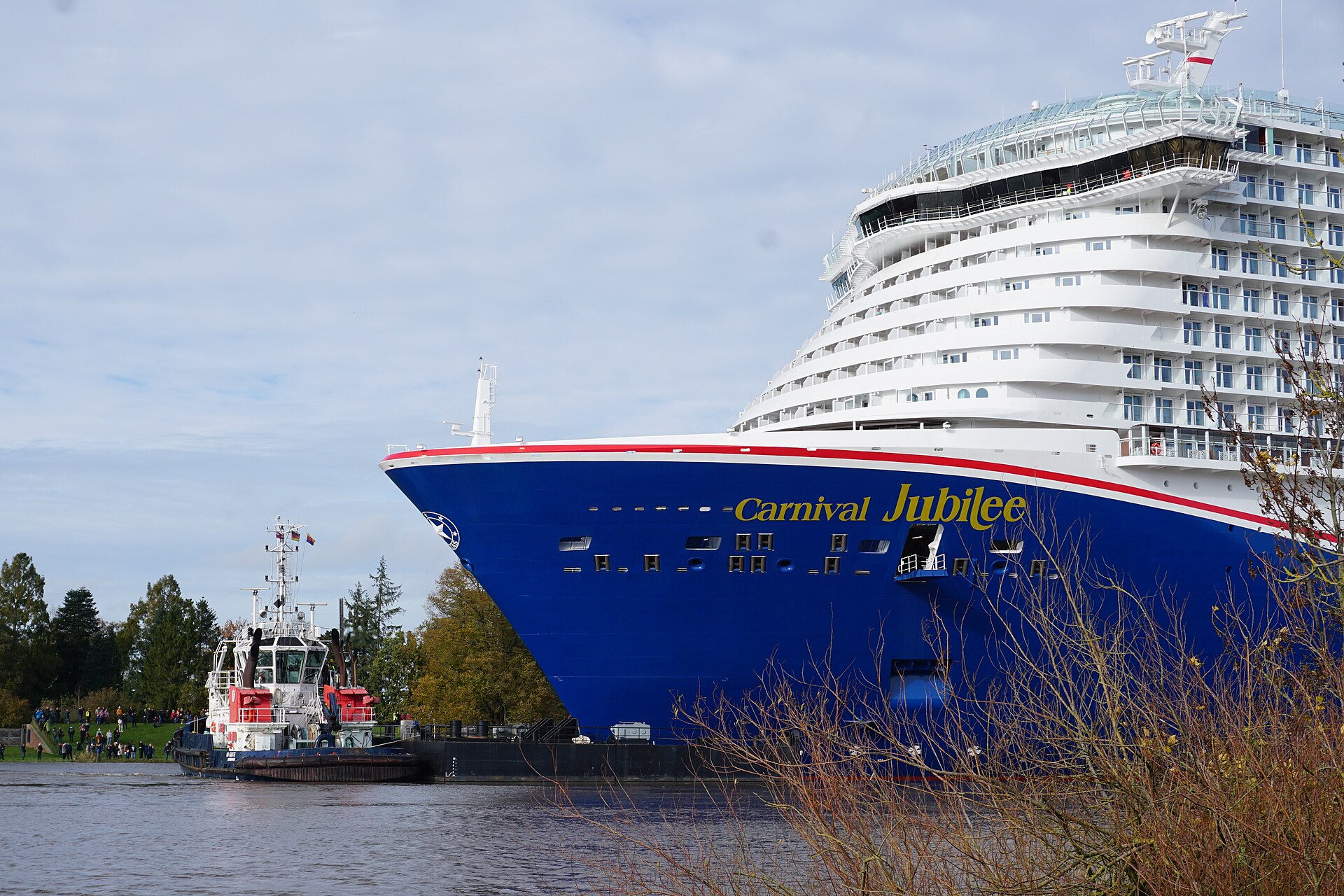 The bright blue bow of Carnival Jubilee with a red-and-white tug alongside on a river in Germany