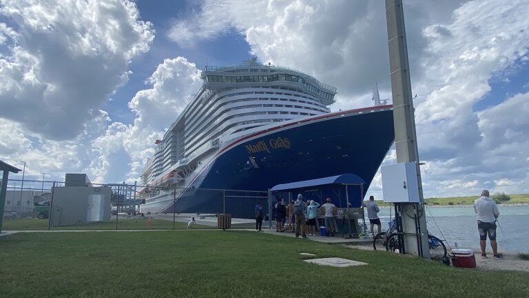 Carnival Mardi Gras docked at Port Canaveral under blue skies and white clouds, with people fishing in the foreground