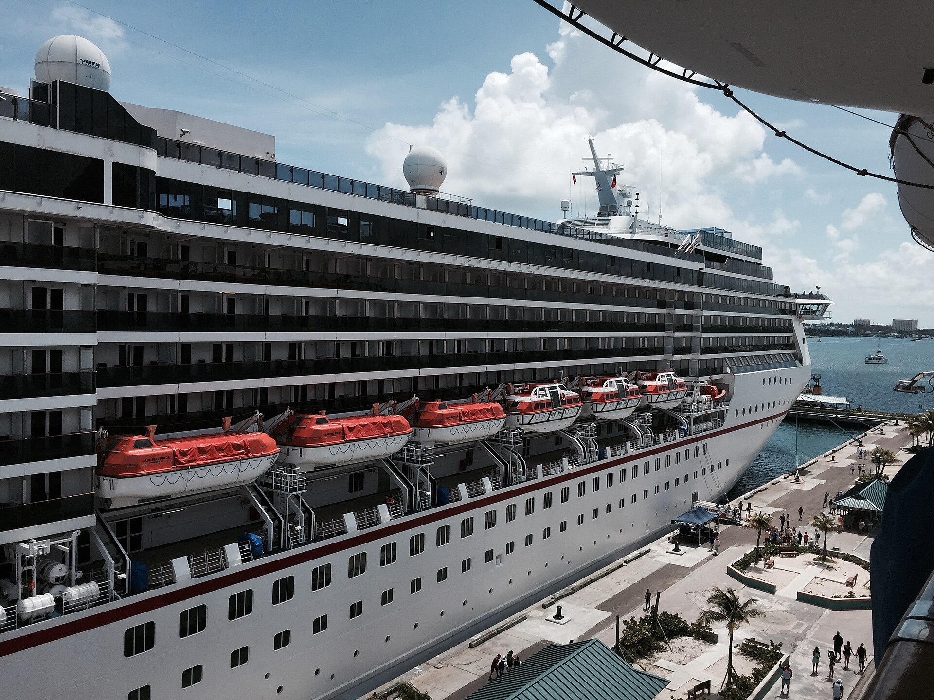 Orange lifeboats lined up along the side of the cruise ship Carnival Pride, docked in Nassau under a blue sky with white clouds