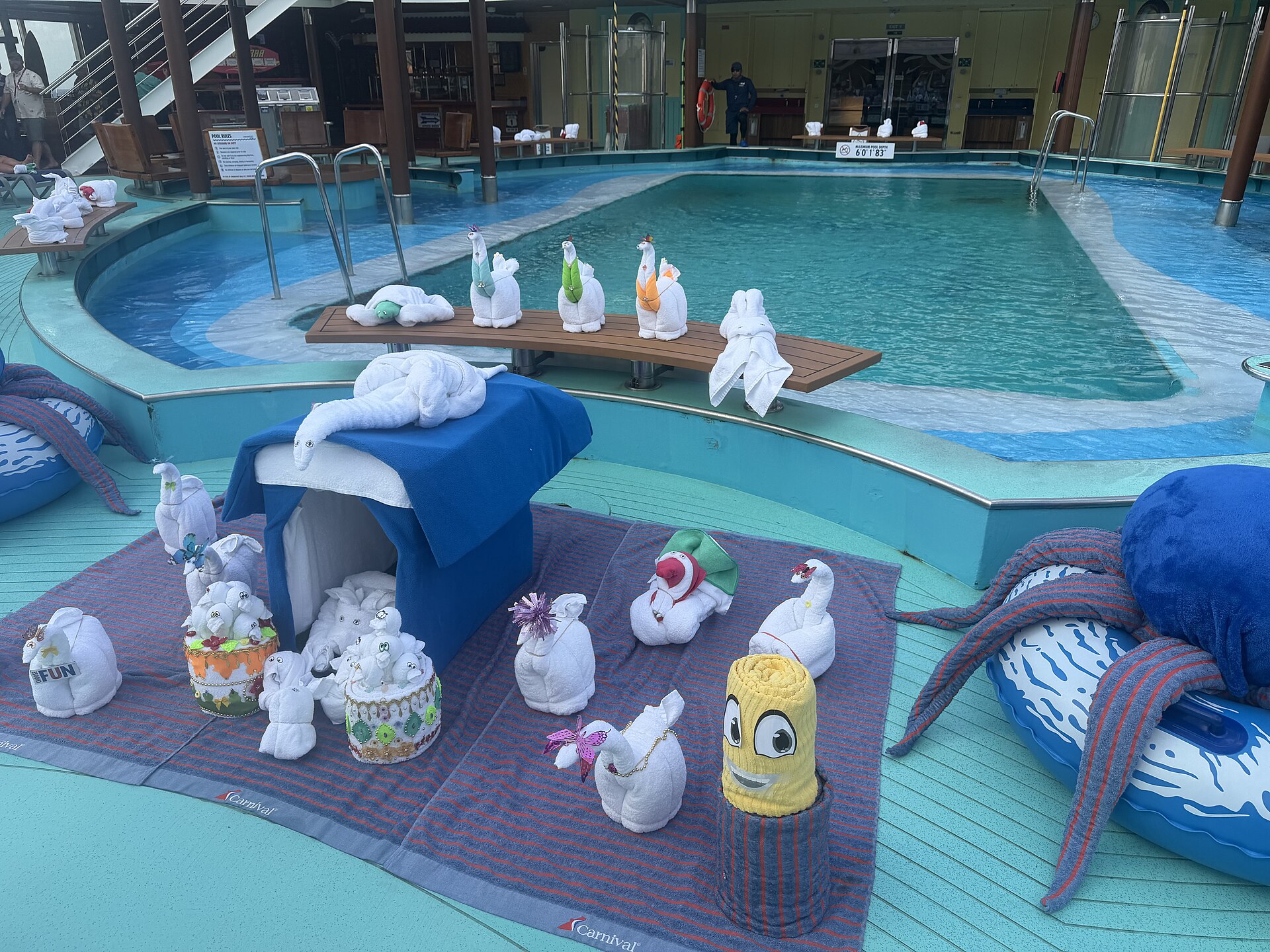 Towel animals including swans, an elephant and a smiling yellow towel figure displayed on Carnival-branded mats beside a turquoise pool