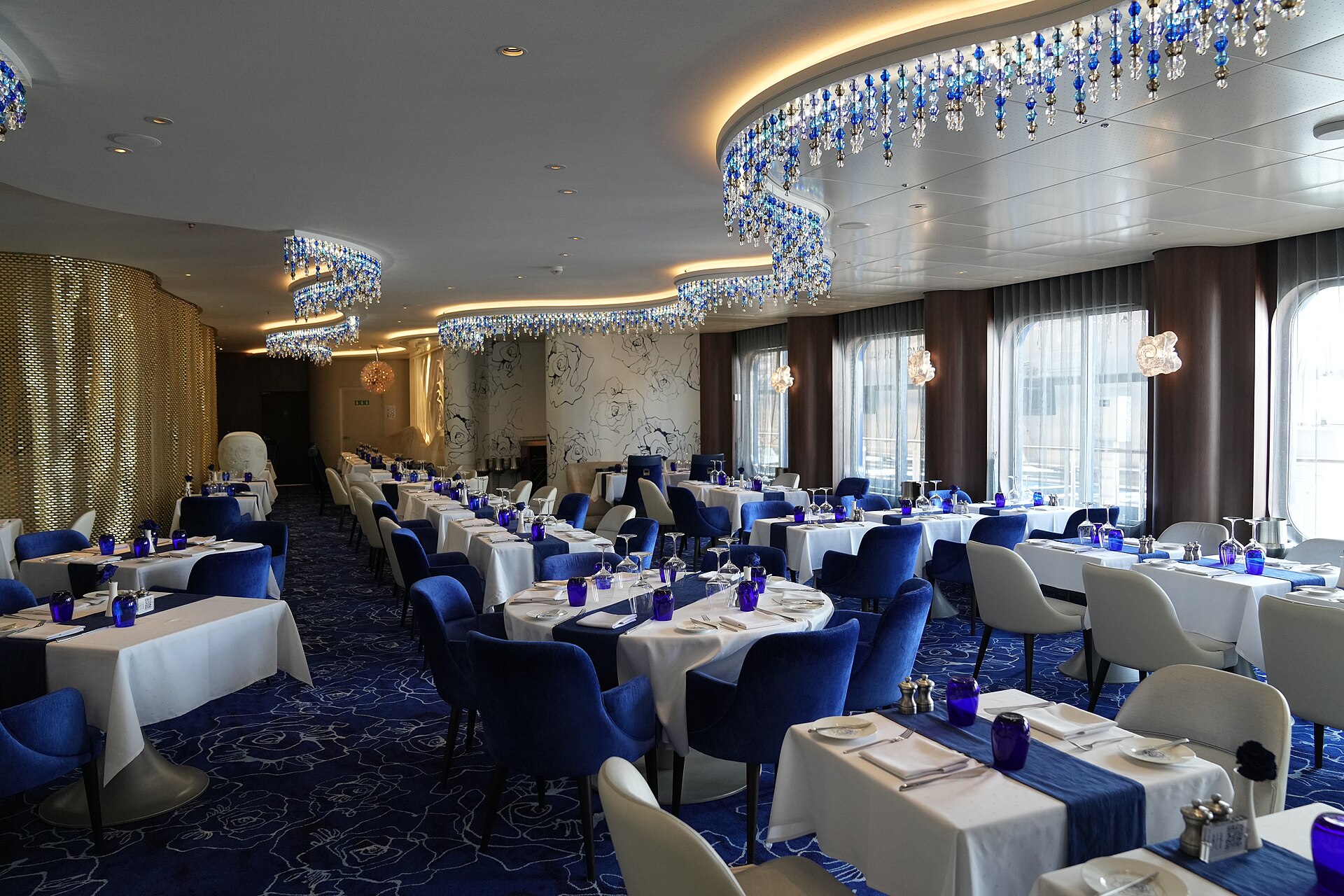 Blu restaurant on Celebrity Apex with bright blue chairs, white tablecloths and blue glass chandeliers beside tall windows