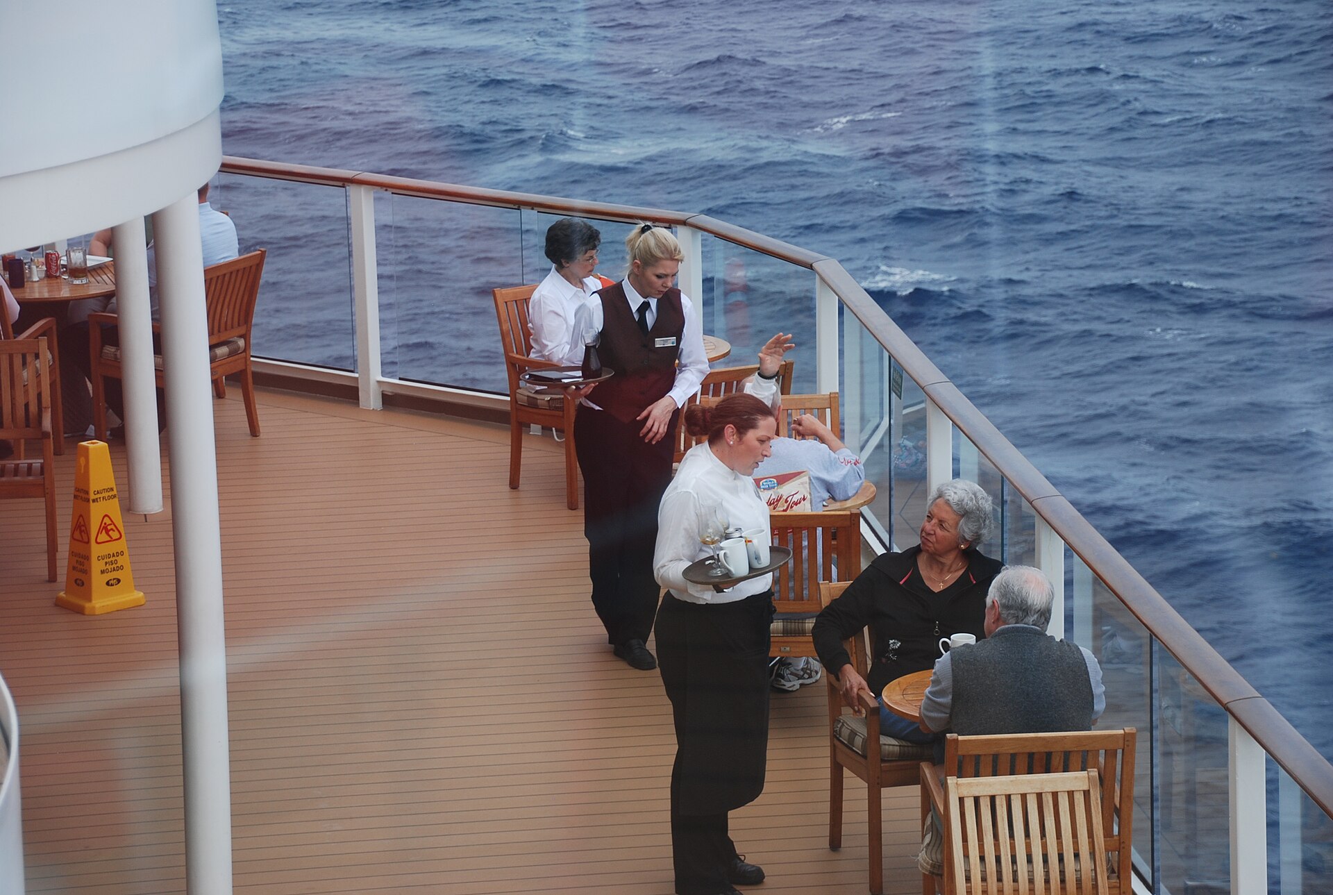 Waitstaff serving drinks to guests at tables on an open deck of Celebrity Equinox above a deep blue sea