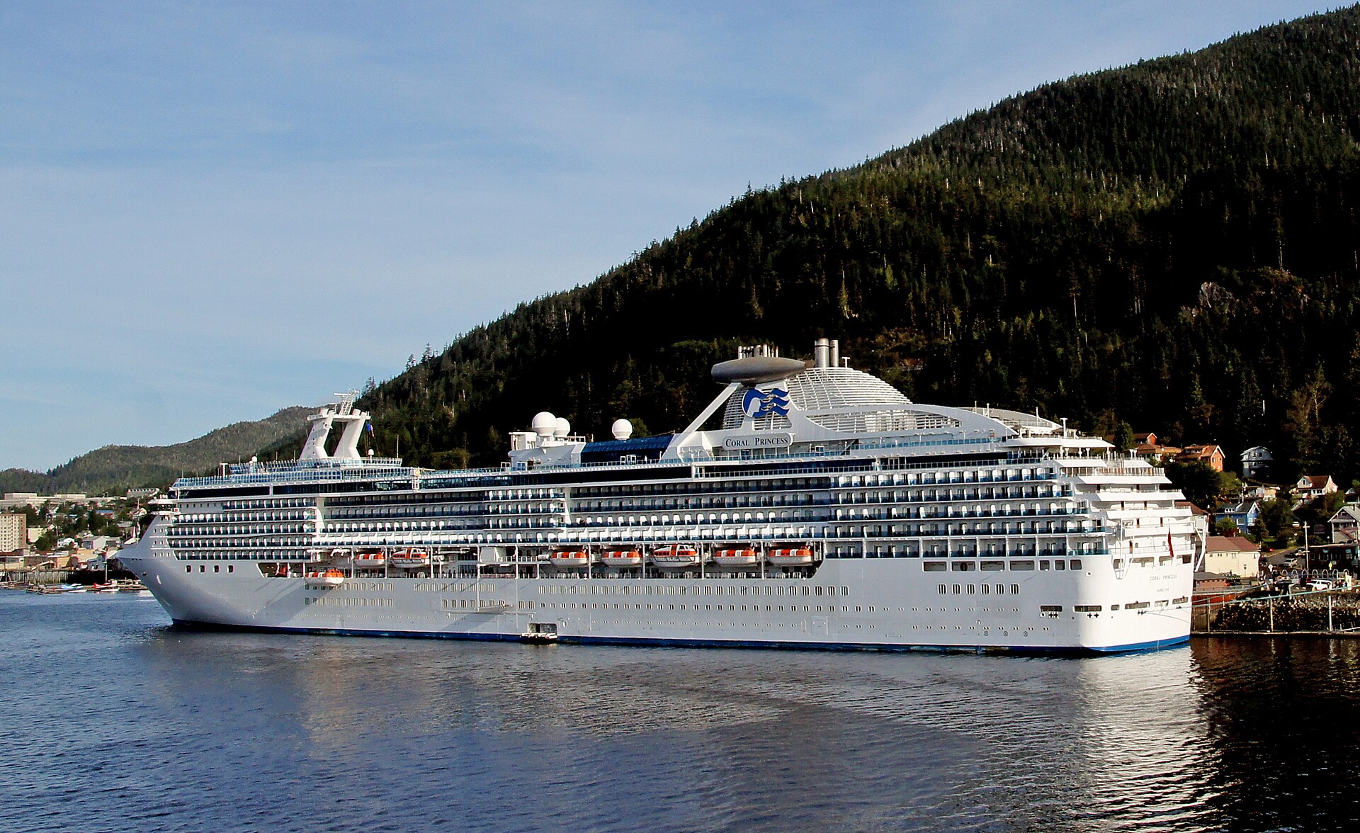 Coral Princess seen side-on in calm water below a forested hillside, with its cabins and lifeboats running from bow to stern