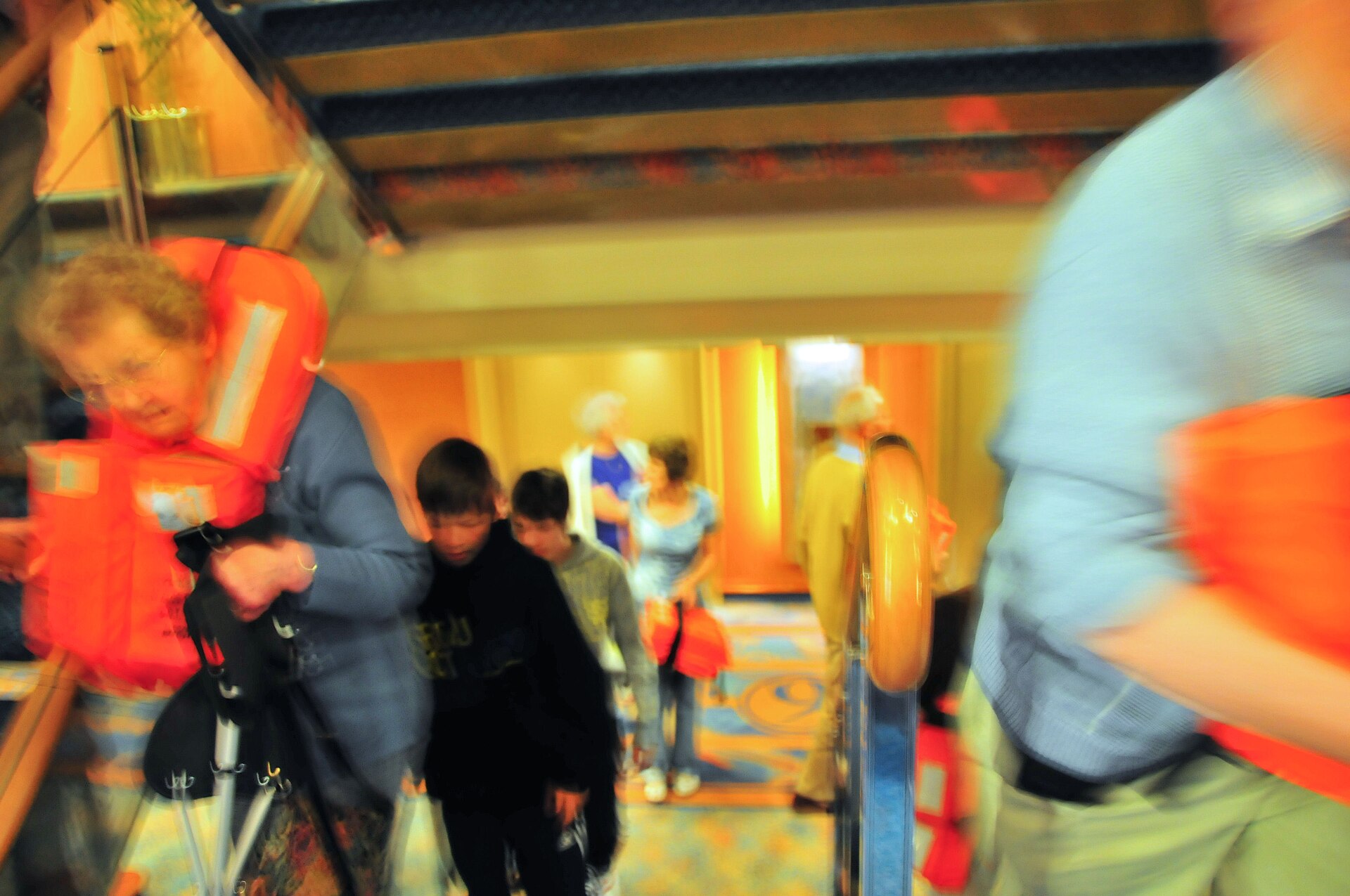Cruise passengers carrying orange life jackets walk to their muster stations during a pre-sailing emergency drill