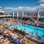 Sunbathers on blue loungers around a bright blue pool on the deck of the cruise ship Britannia under a sunny sky