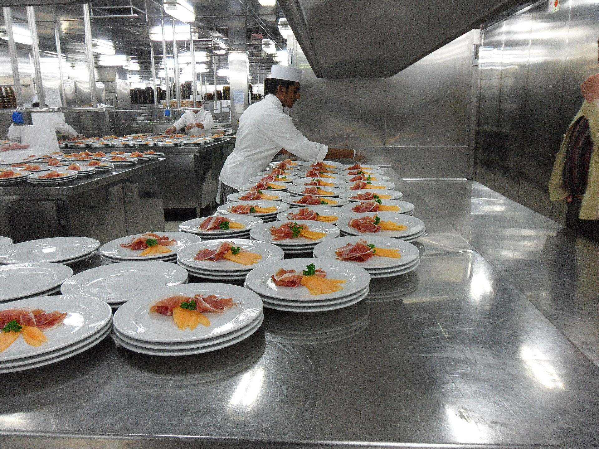 A chef plating rows of starters on a stainless steel counter in a cruise ship galley