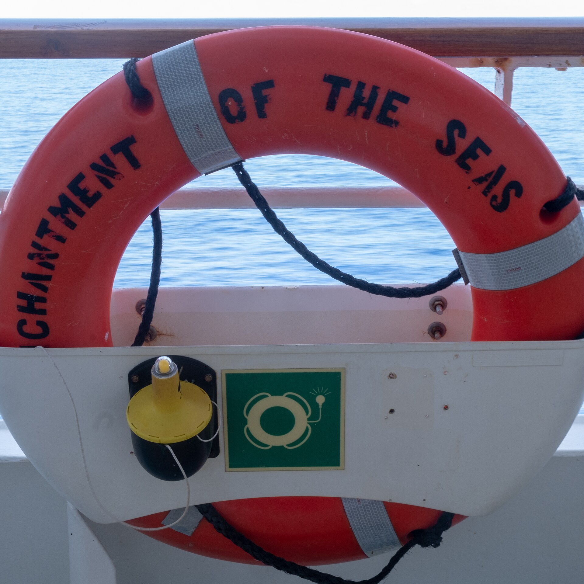 An orange lifebuoy on the rail of Royal Caribbean's Enchantment of the Seas, with its yellow self-igniting light mounted beside it and blue sea behind