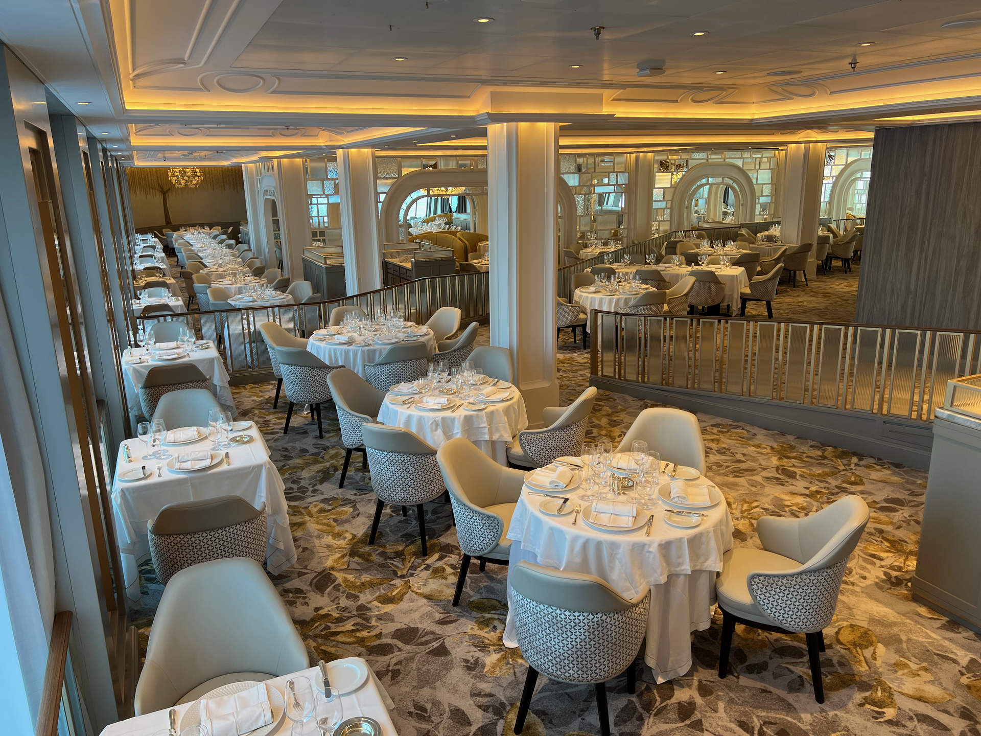 A bright cruise ship main dining room with white tablecloths, cream chairs and a patterned carpet, set for dinner