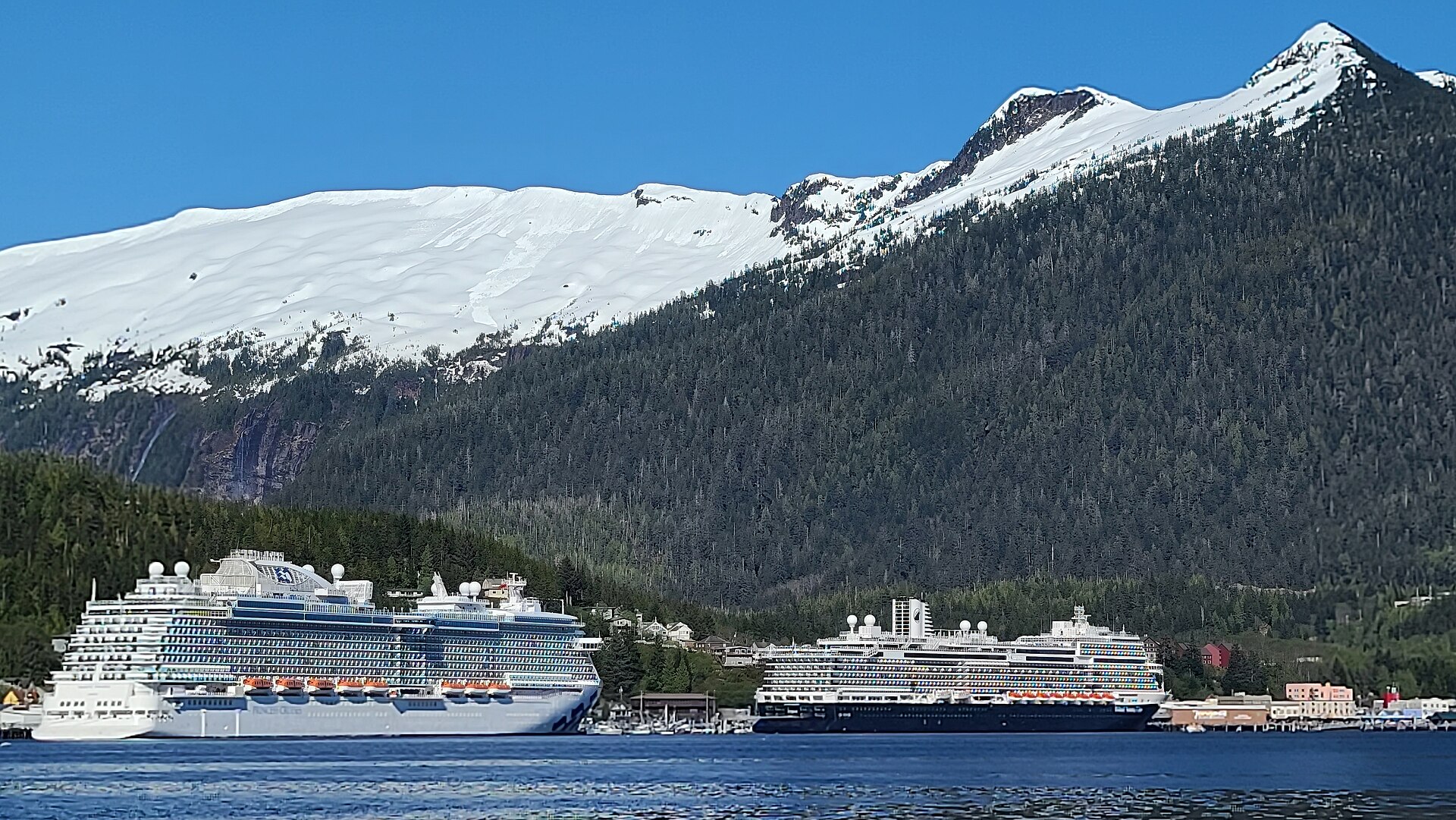 Two large cruise ships docked along the Ketchikan waterfront under snowy, forested mountains and a clear blue sky