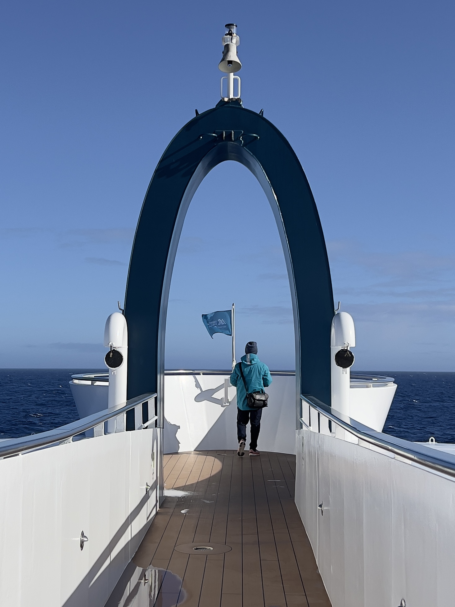 A passenger in a turquoise jacket on the bow of an expedition ship under a dark blue arch, with deep blue sea and sky ahead in the Drake Passage