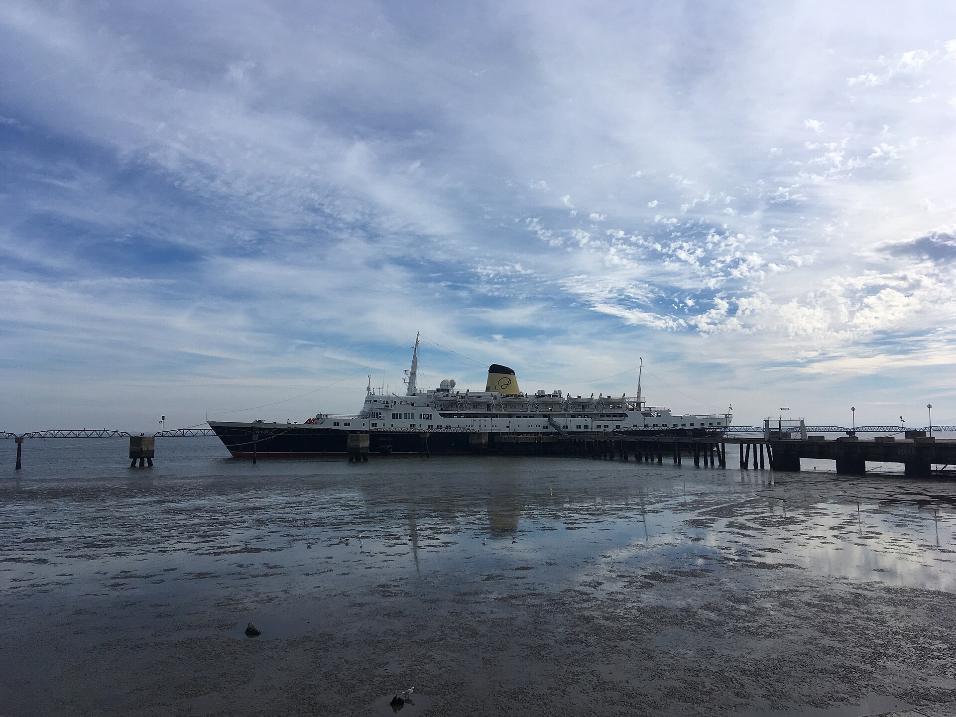 Funchal moored at a long pier on the Tagus River at low tide, with a dramatic blue sky and streaked white clouds