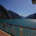 Turquoise, glassy water between steep forested mountains and snowy peaks in Alaska's Inside Passage, seen from a cruise ship deck