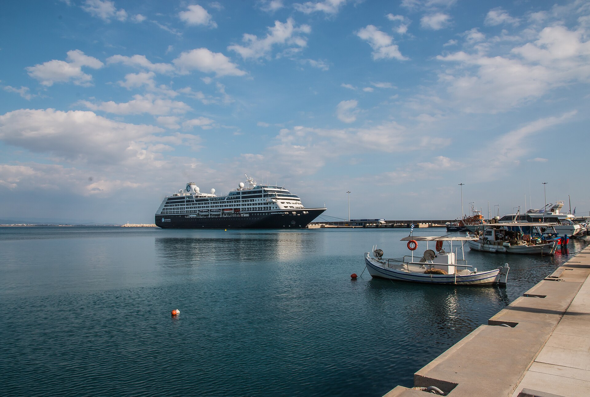A cruise ship anchored off the harbour at Katakolo, Greece, with fishing boats moored along the quay under a blue sky