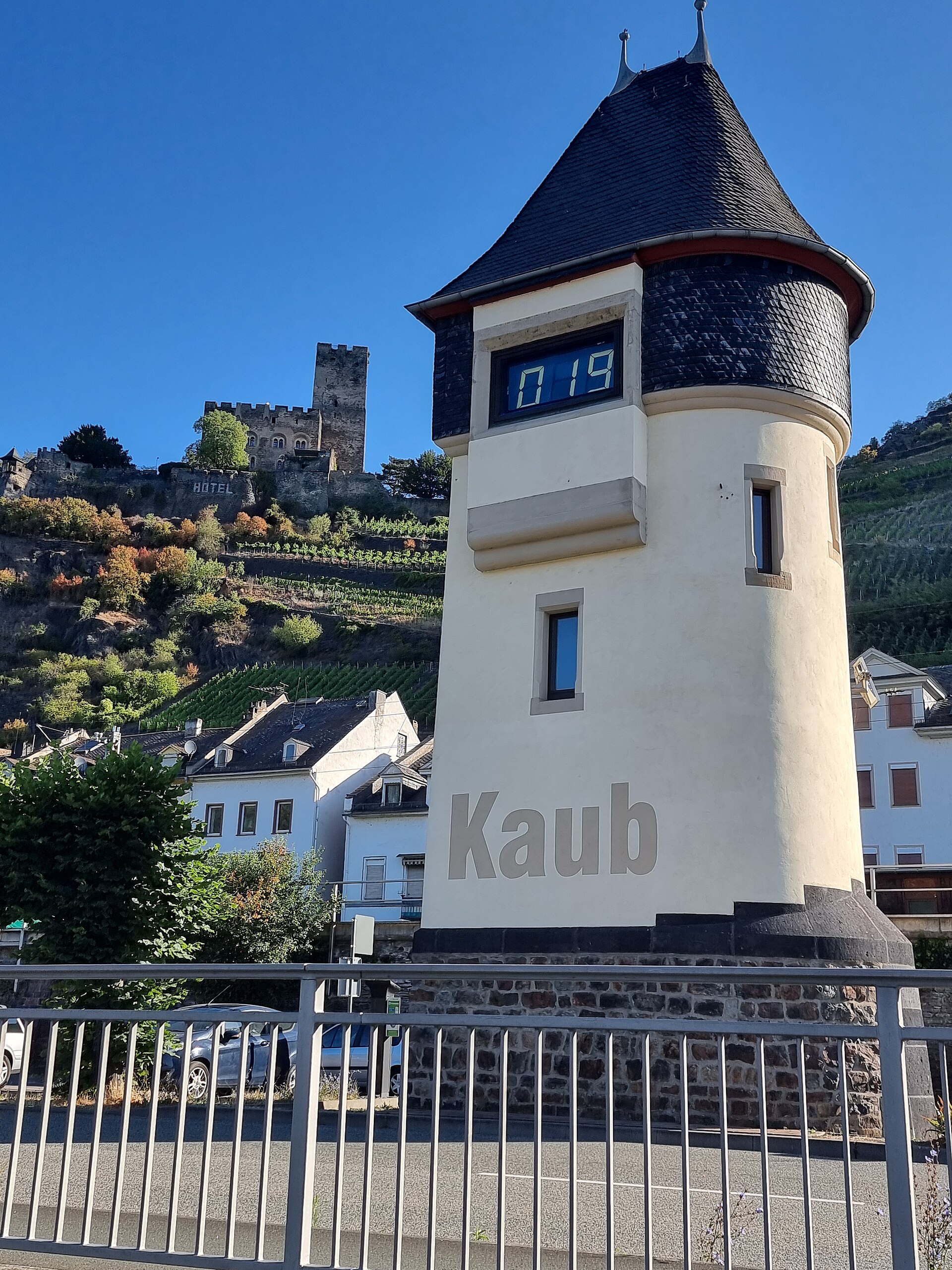 The white Kaub gauge tower on the Rhine with its digital display reading 019, vineyards and a hilltop castle behind under a blue sky