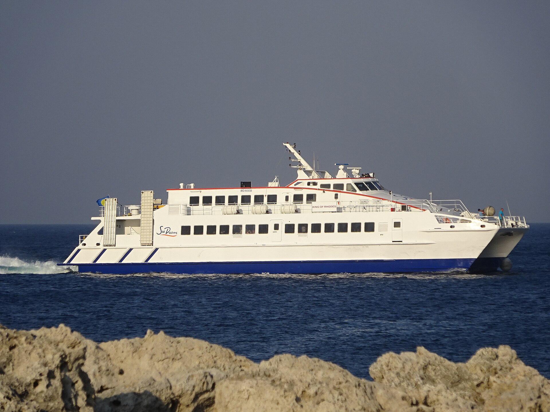 The catamaran King of Rhodes speeding across deep blue water past a rocky shore
