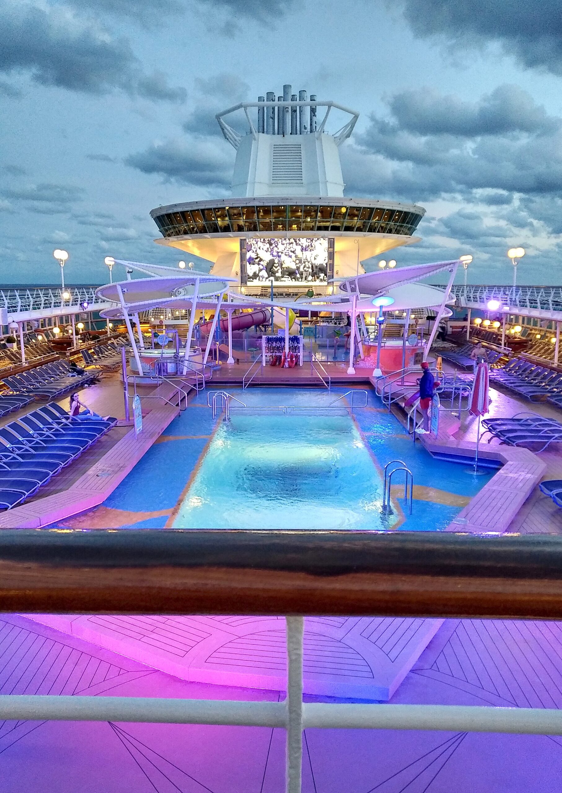 Majesty of the Seas pool deck at dusk, with a blue pool, rows of loungers, pink deck lighting and an outdoor movie screen under the funnel