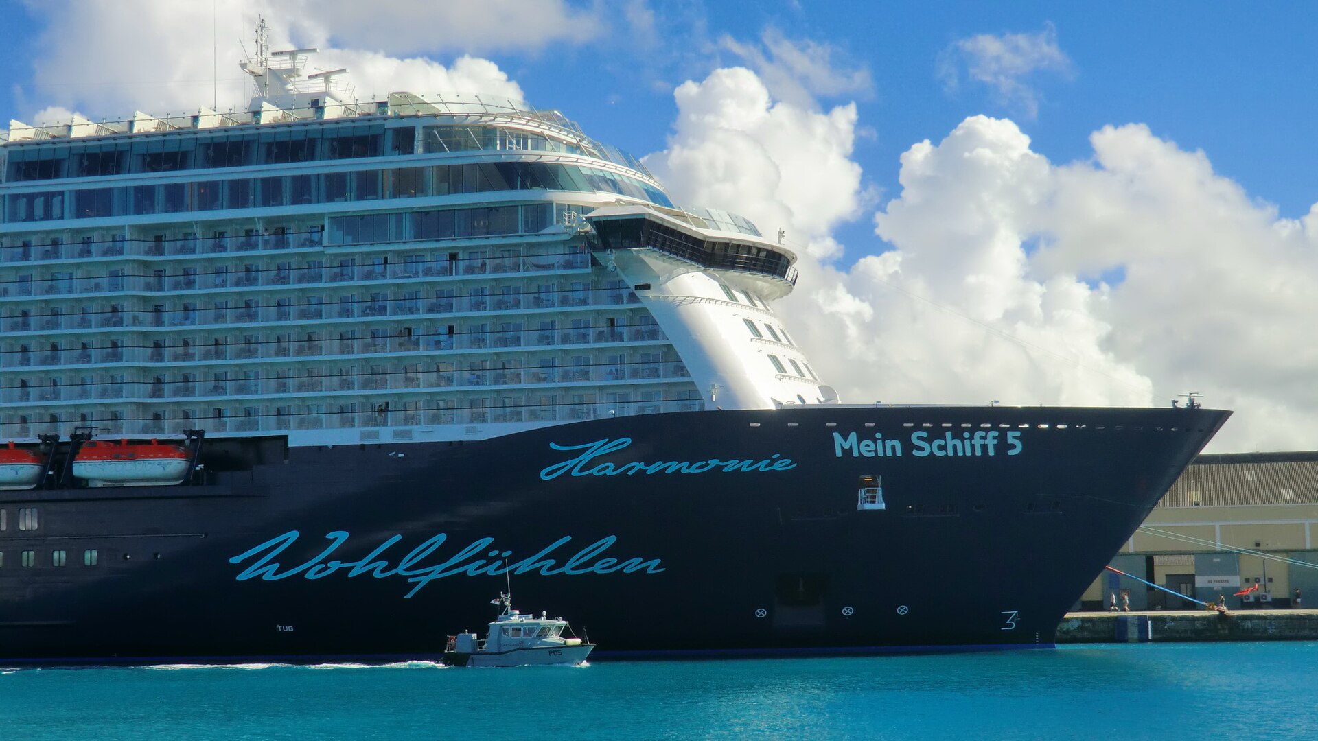 The dark blue hull of Mein Schiff 5 docked in bright turquoise water at Bridgetown, Barbados, with a small pilot boat alongside