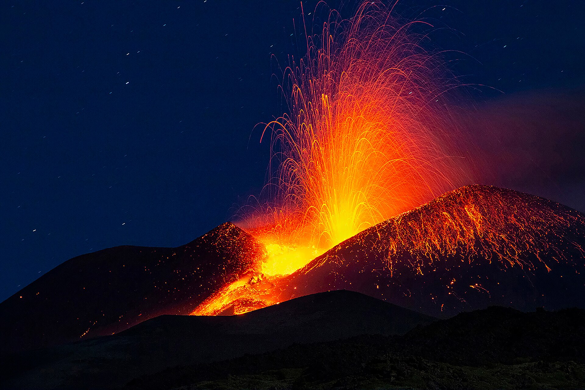 Mount Etna erupting at night, with a fountain of orange lava arcing over a dark cone under a starry blue sky