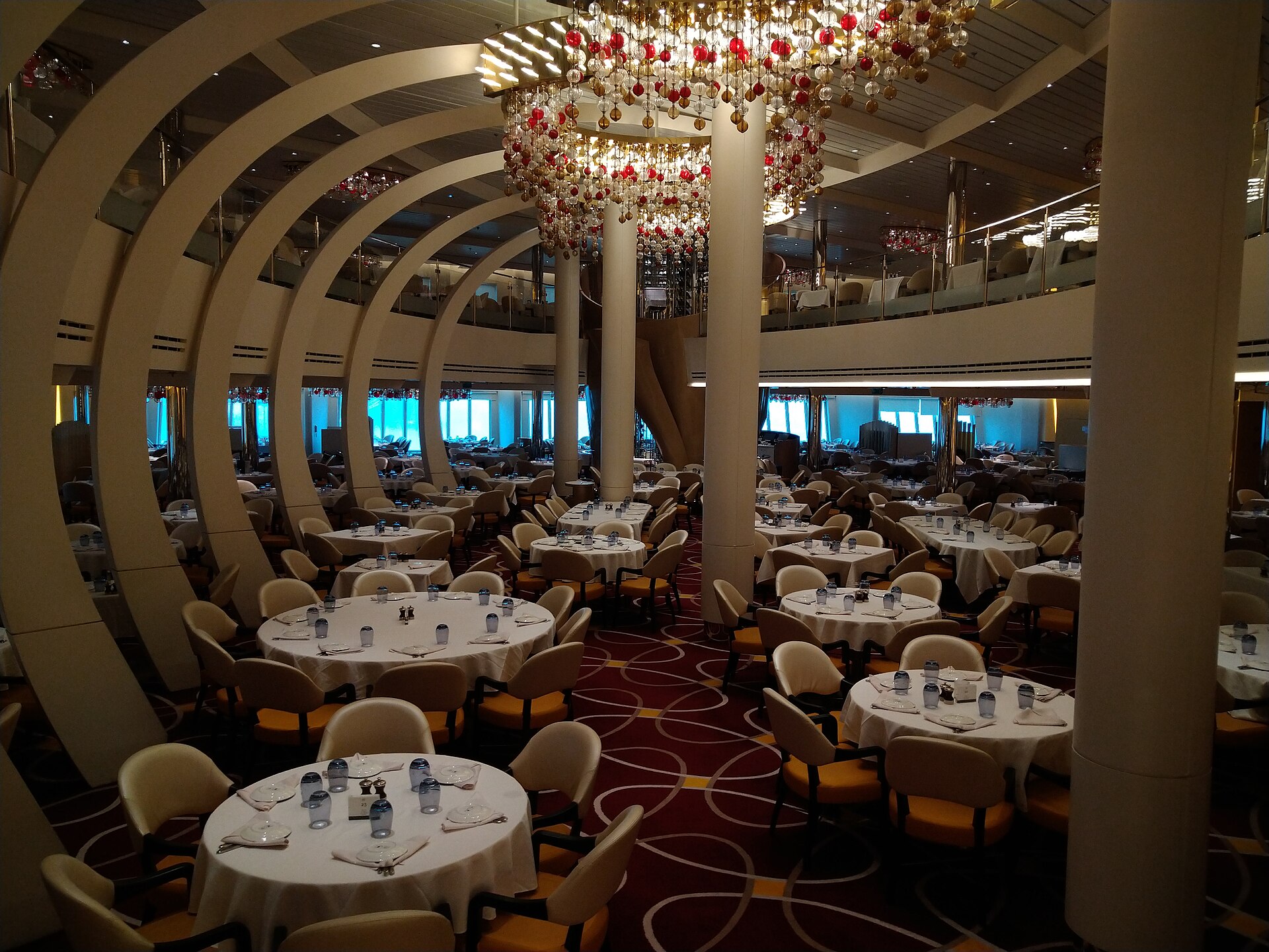 The two-level main dining room on Holland America's Nieuw Statendam, with round tables, cream and orange chairs and a red glass chandelier