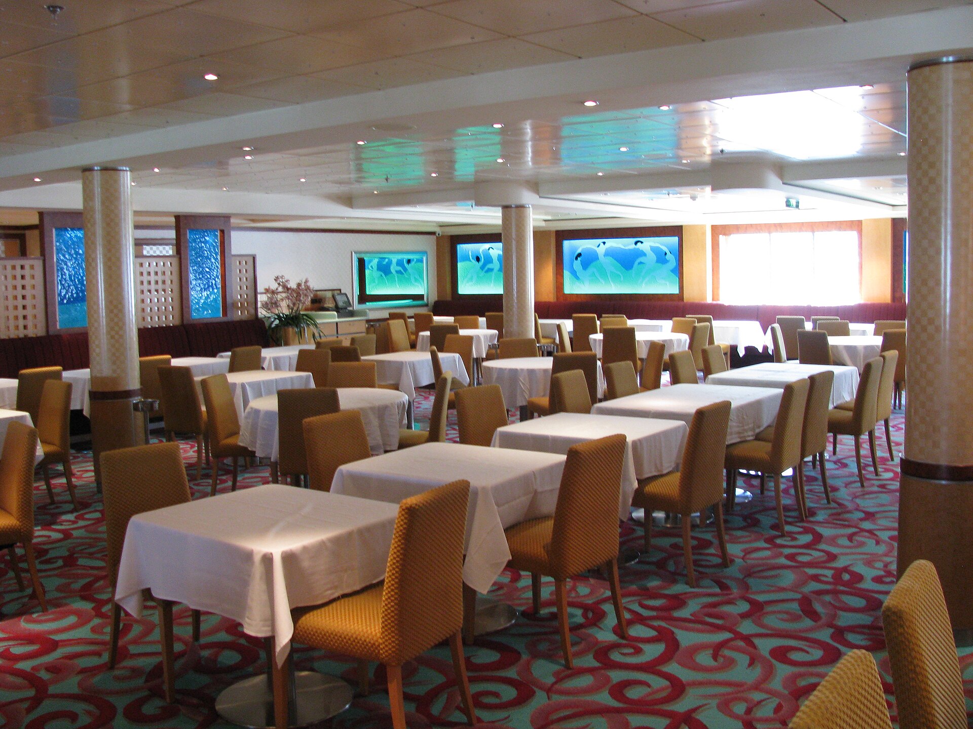 The Aqua dining room on Norwegian Dawn with white tablecloths, gold chairs, a patterned red carpet and bright blue wall panels